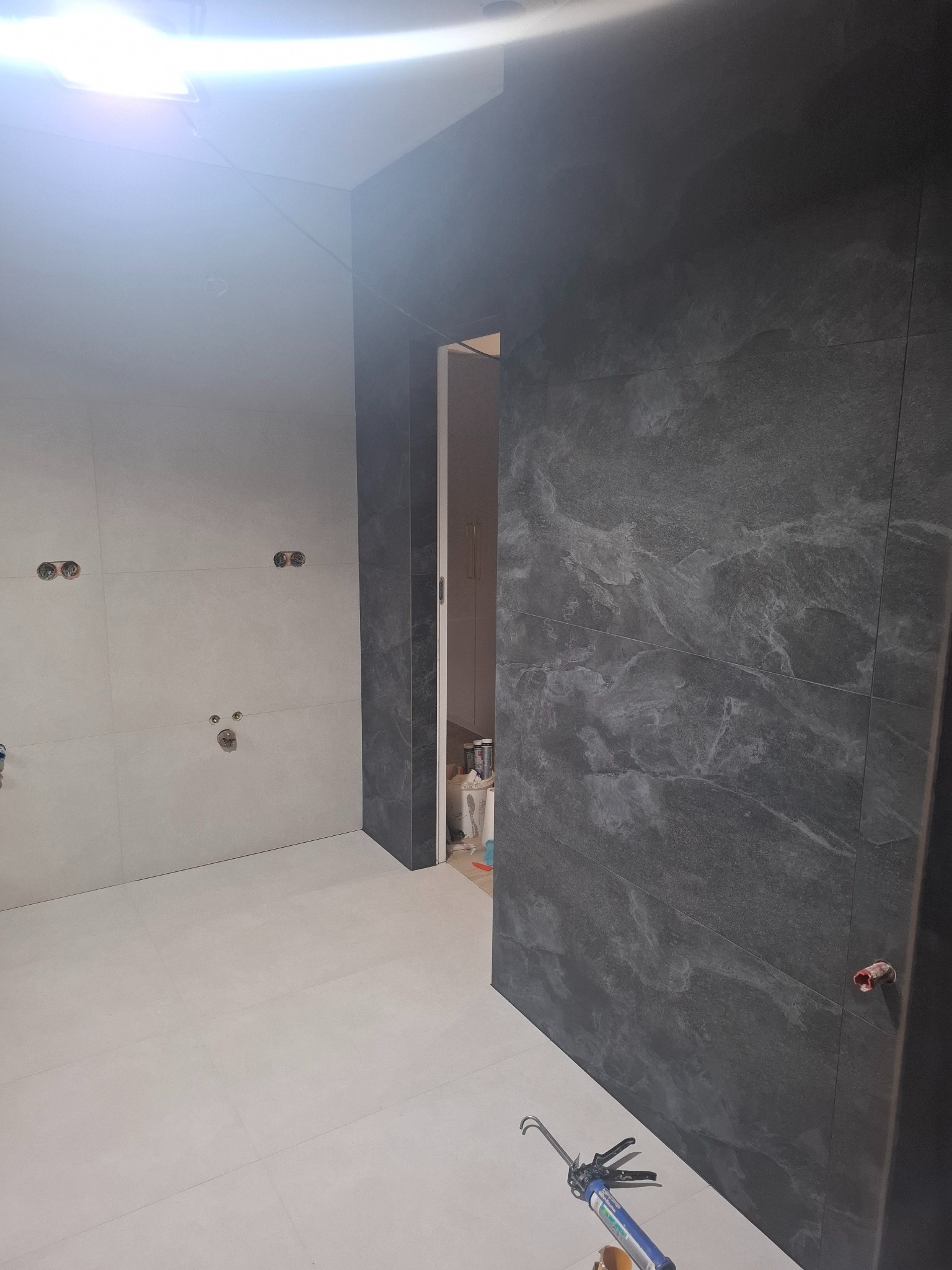 Wnętrze łazienki w trakcie prac wykończeniowych: ułożone szare płytki imitujące kamień na ścianie i podłodze, widoczne otwory na instalacje, narzędzie do silikonu leżące na podłodze, w tle otwarte...