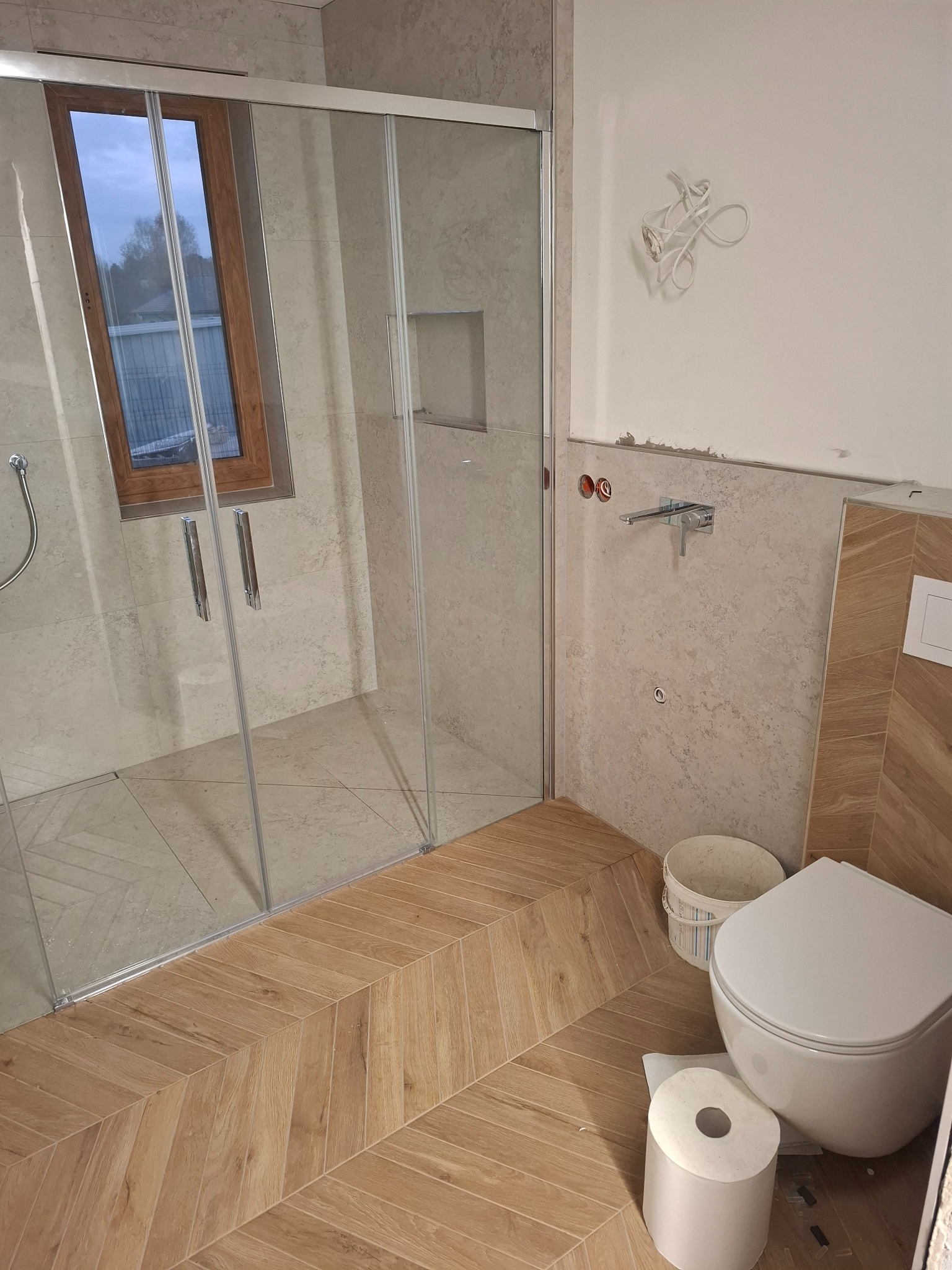 Wnętrze łazienki w trakcie prac wykończeniowych: kabina prysznicowa ze szklanymi drzwiami, fragment ściany z zamontowaną baterią, toaleta, wiadro i rolka papieru toaletowego na podłodze...