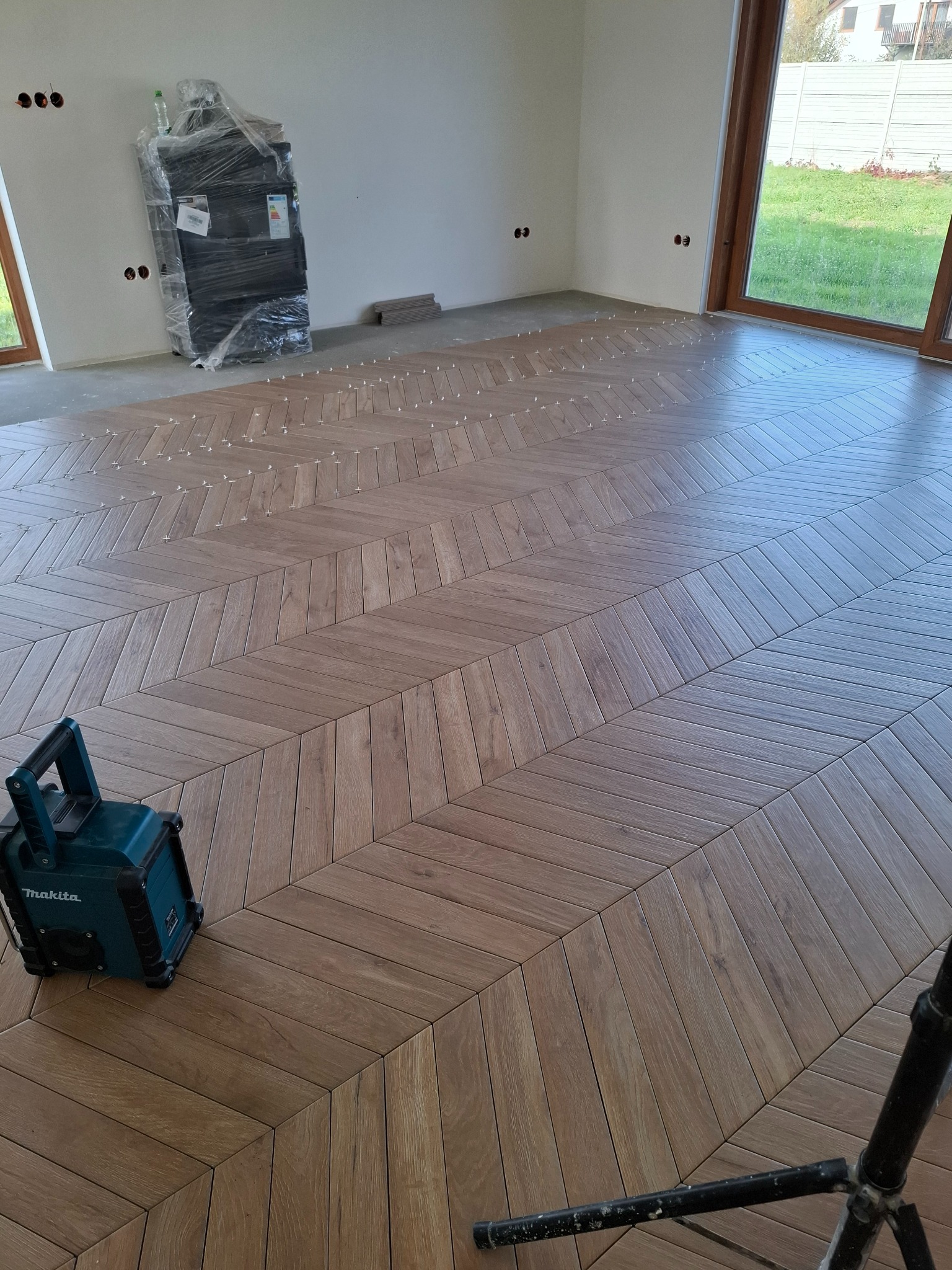 Układanie drewnopodobnych płytek w jodełkę w przestronnym, jasnym wnętrzu; widoczne krzyżyki dystansowe, urządzenie Makita i zapakowane elementy wyposażenia.