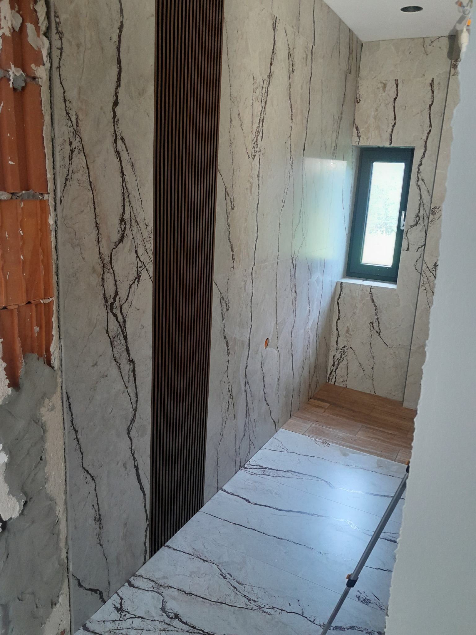 Wnętrze łazienki w trakcie remontu: ściany wyłożone płytkami imitującymi marmur z brązowymi żyłami, dekoracyjny panel z pionowych drewnianych listew, fragment odsłoniętej cegły, małe okno z ciemną...