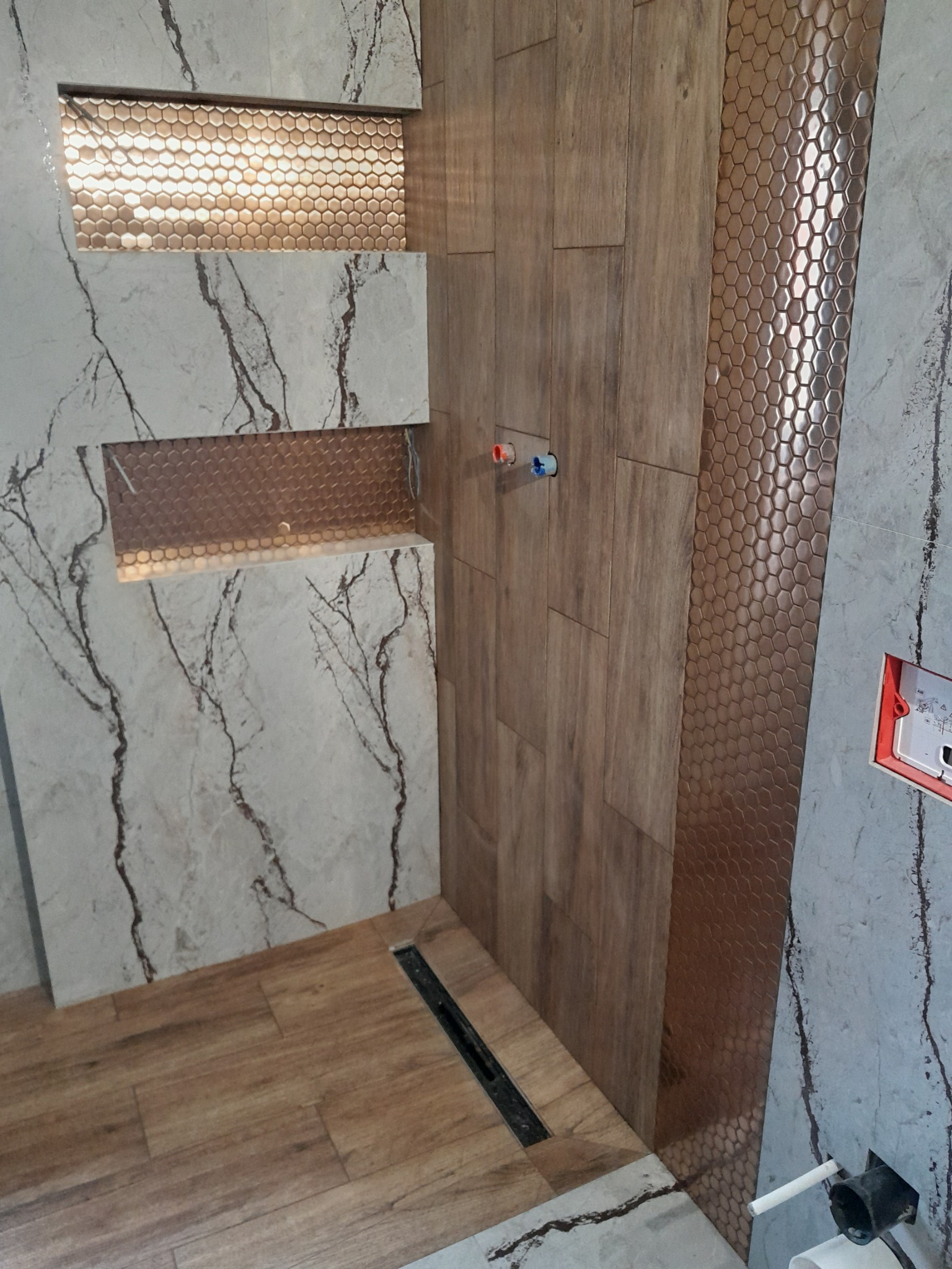 Nowoczesna aranżacja łazienki w Grywałdzie: połączenie płytek imitujących drewno, marmuru i mozaiki heksagonalnej, z podświetlanymi wnękami i odpływem liniowym w podłodze.