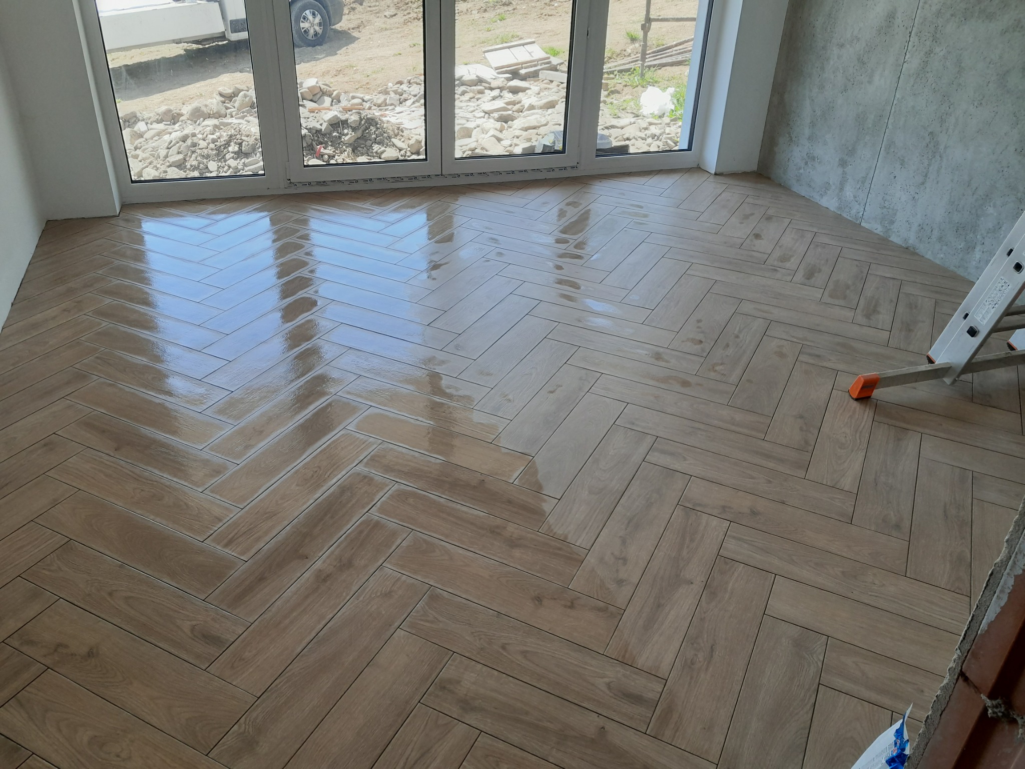 Świeżo ułożona, błyszcząca podłoga z płytek imitujących drewno ułożonych w jodełkę, widok na okno z widokiem na plac budowy i fragment drabiny malarskiej.
