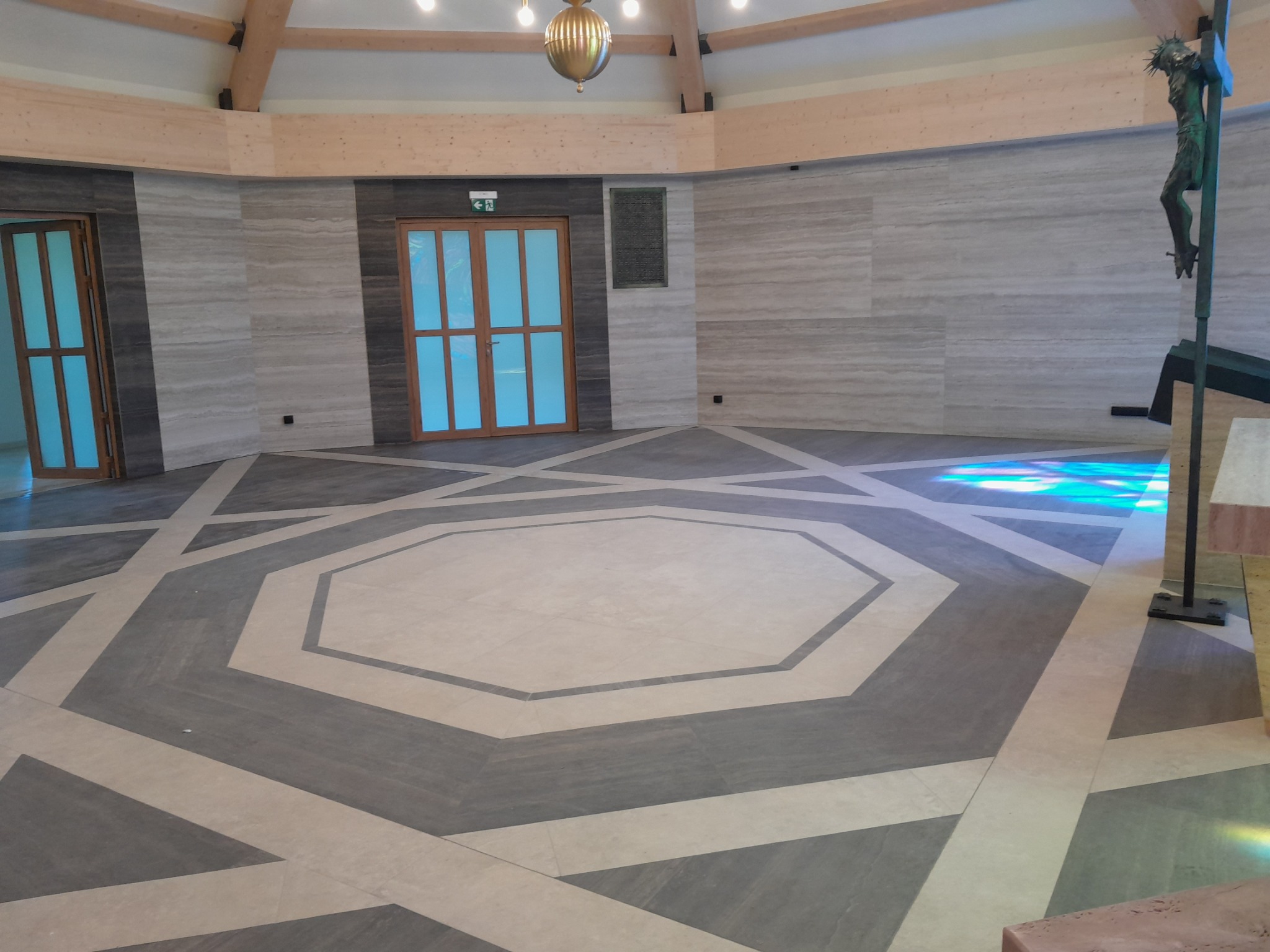 Wnętrze kościoła z geometrycznym wzorem na podłodze z płytek, jasne ściany z efektem marmuru, drewniane belki stropowe i rzeźba ukrzyżowanego Chrystusa po prawej.