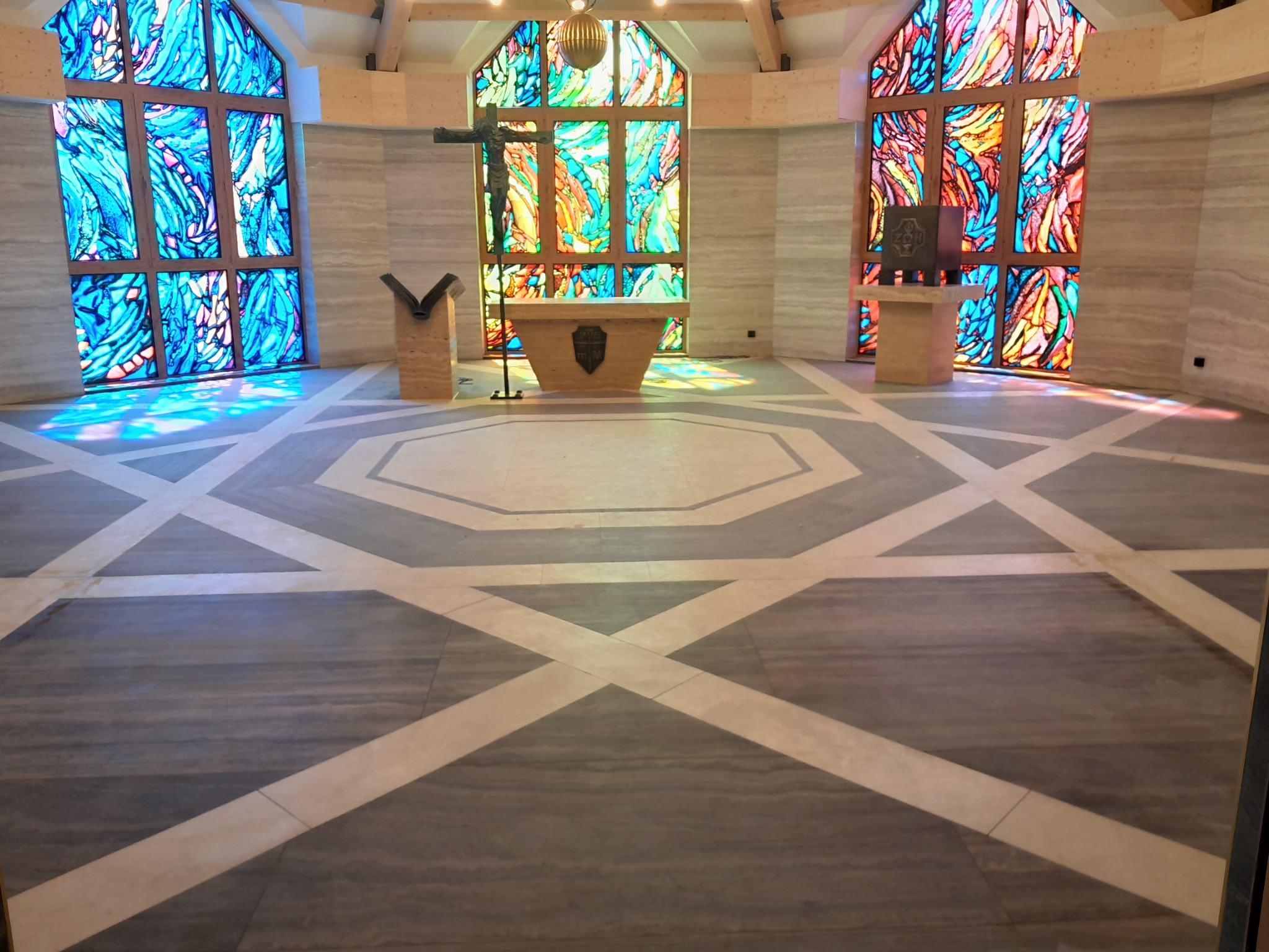 Wnętrze kaplicy z geometryczną posadzką w odcieniach szarości i beżu, marmurowe ściany oraz witraże w oknach, rzucające kolorowe refleksy świetlne na podłogę.