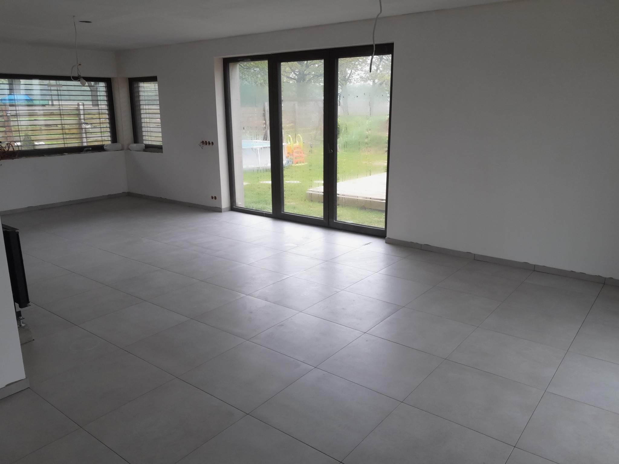 Wnętrze pustego pokoju z szarymi, kwadratowymi płytkami podłogowymi, dużym oknem balkonowym z widokiem na ogród z placem zabaw i dwoma mniejszymi oknami z żaluzjami, ściany pomalowane na biało...