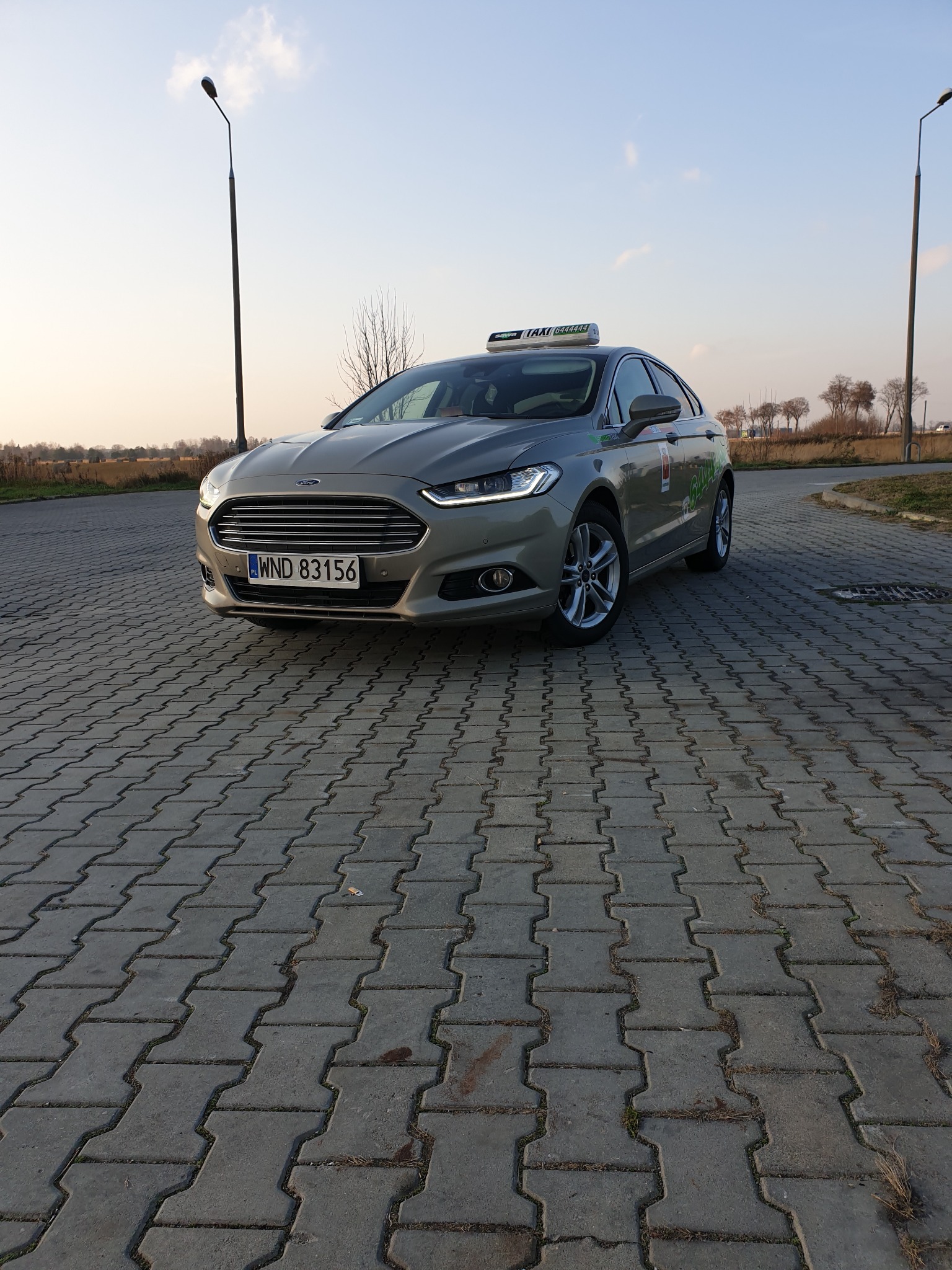 Szary samochód osobowy z oznaczeniem Taxi Sava 6444444 na dachu, zaparkowany na brukowanej nawierzchni, widok z przodu, tablica rejestracyjna WND 83156.