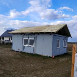 Kontener biurowy z drewnianą konstrukcją dachu w trakcie budowy na tle pochmurnego nieba i sąsiedniego domu.