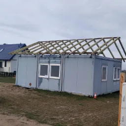 Kontener biurowy z drewnianą konstrukcją przygotowaną pod dach, widoczny w trakcie budowy na zewnątrz.