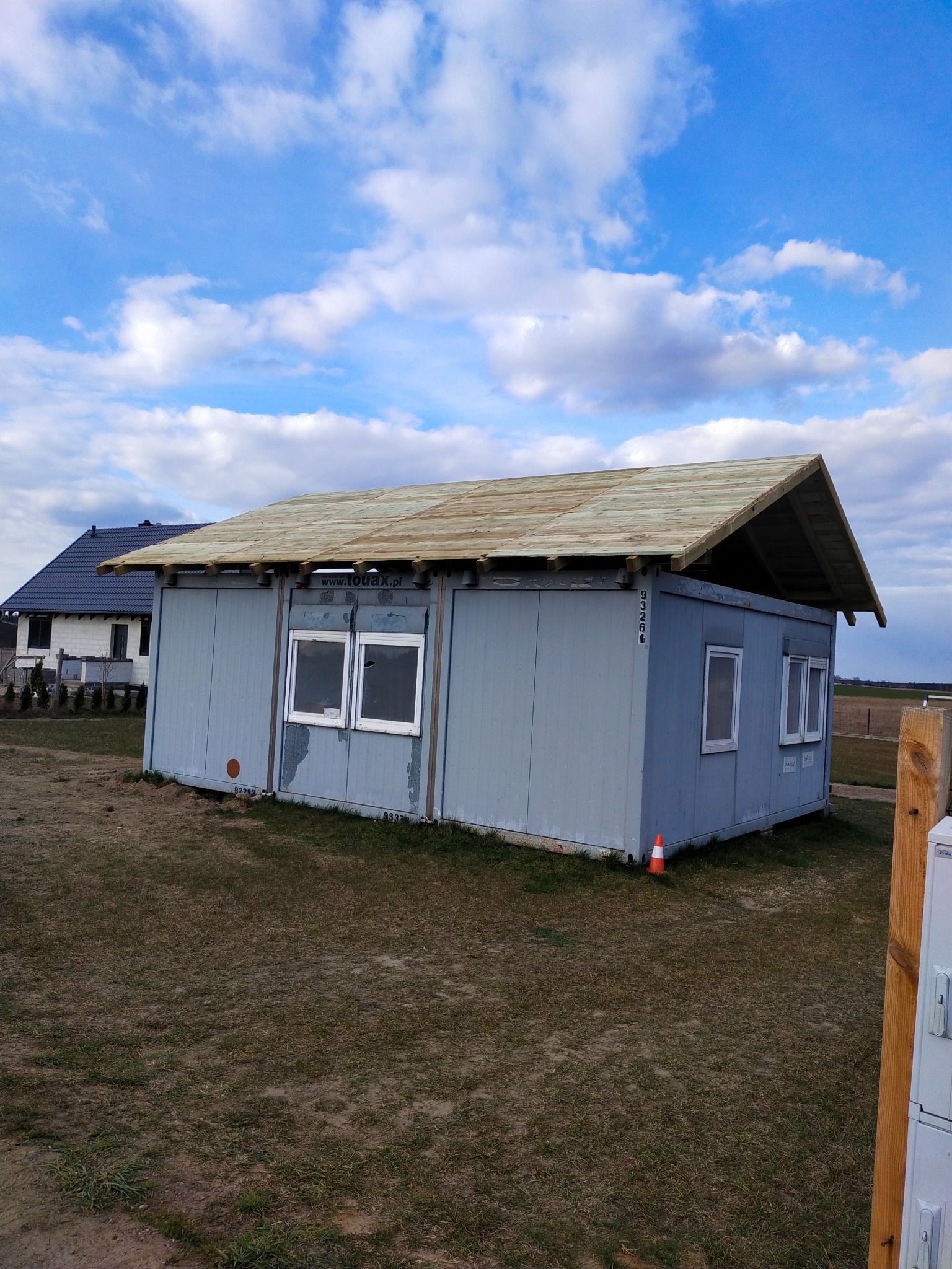 Kontener biurowy z drewnianą konstrukcją dachu w trakcie budowy na tle pochmurnego nieba i sąsiedniego domu.