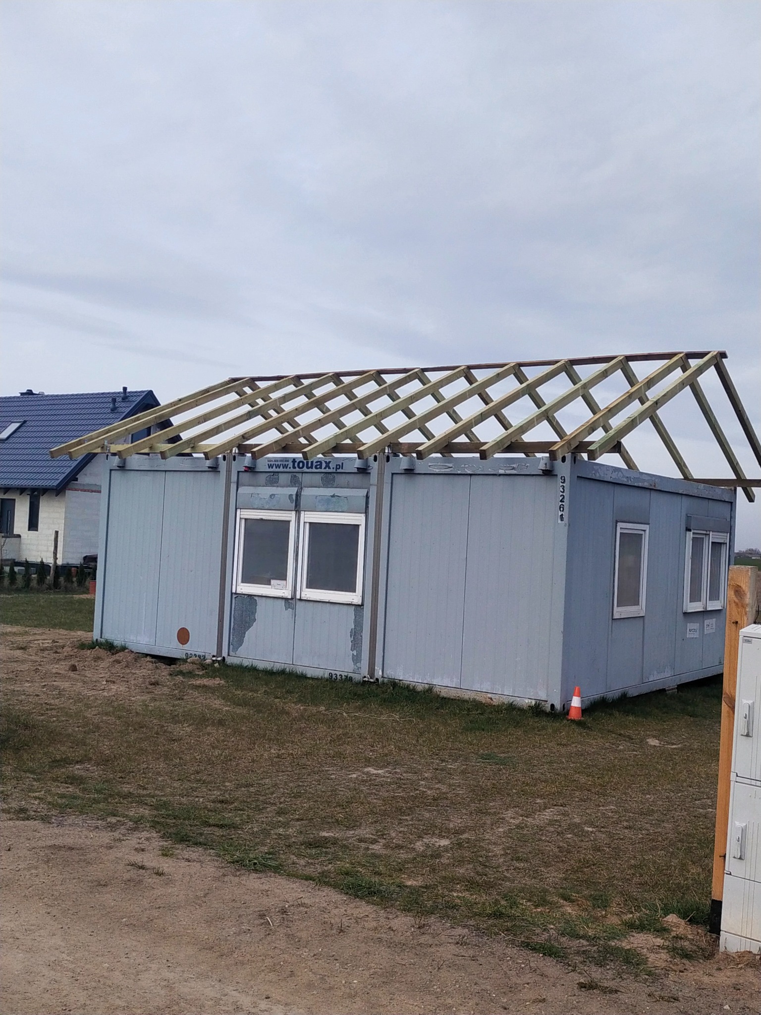 Kontener biurowy z drewnianą konstrukcją przygotowaną pod dach, widoczny w trakcie budowy na zewnątrz.