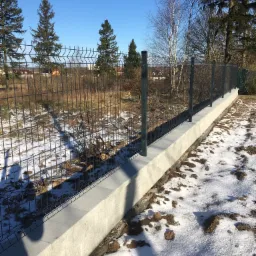 Nowo postawione ogrodzenie panelowe na betonowej podmurówce w zimowym krajobrazie z resztkami śniegu na ziemi.