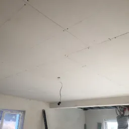 Montaż sufitu podwieszanego z płyty karton-gips, widoczne linie wyznaczające punkty mocowania, przewody elektryczne wystające z sufitu, pracownik w czerwonych rękawicach instaluje konstrukcję.