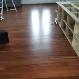 Świeżo wycyklinowana, lakierowana podłoga z egzotycznego drewna, widoczny fragment regału z jasnego drewna, drabina malarska i czarny puf.