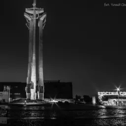 Czarno-białe nocne zdjęcie Pomnika Poległych Stoczniowców 1970 w Gdańsku, z napisem 'Stocznia Gdańska' w tle, widok z brukowanego placu.
