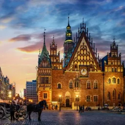 Rynek we Wrocławiu o zmierzchu: iluminowany Ratusz, dorożka konna na pierwszym planie, restauracje z parasolami w tle, malownicze niebo.