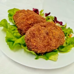 Trzy kotlety panierowane, ułożone na białym talerzu z zieloną i czerwoną sałatą, na tle białego obrusu.