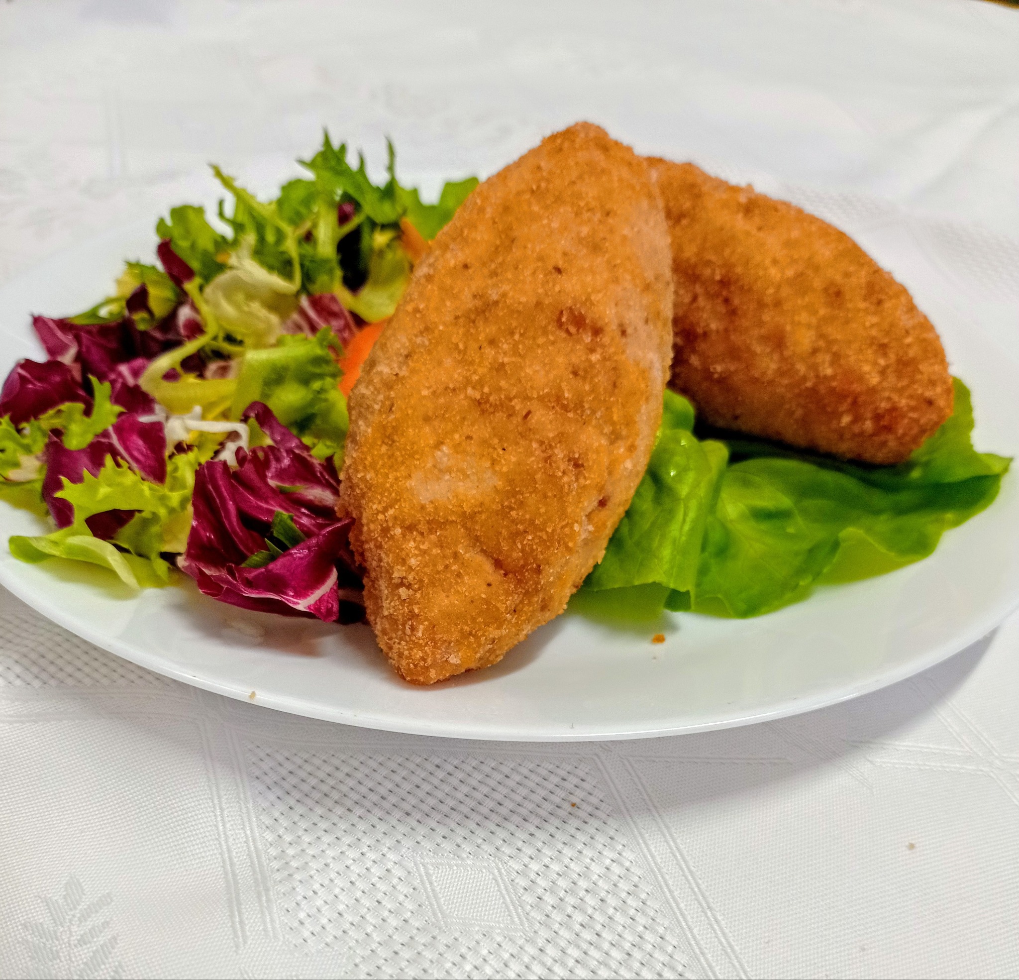 Dwa krokiety w złocistej panierce, podane na liściach sałaty i mieszance sałat na białym, okrągłym talerzu, na białym obrusie z delikatnym wzorem.