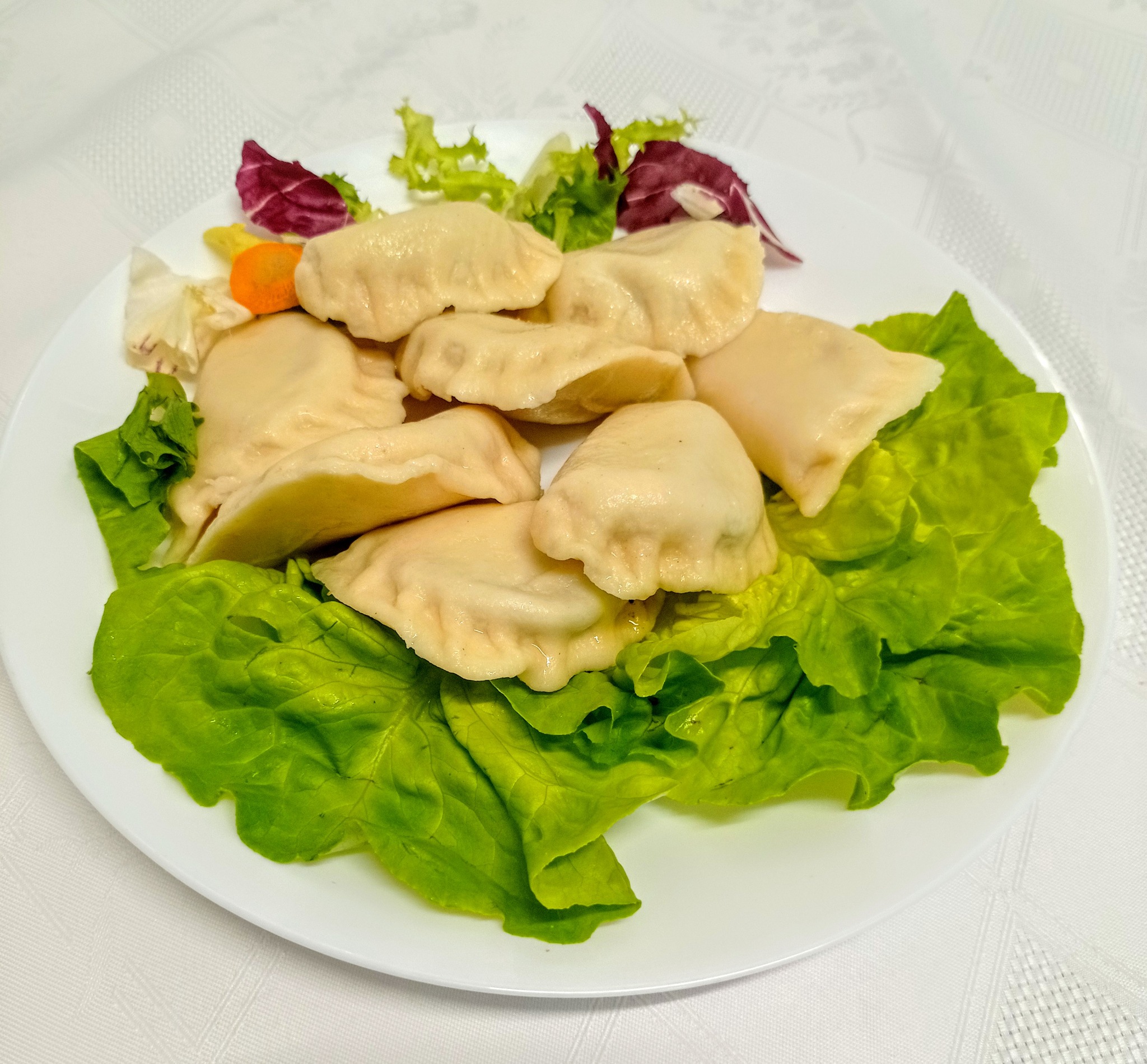 Porcja gotowanych pierogów ułożona na talerzu z zieloną sałatą i dekoracją z sałat mieszanych oraz plasterka marchewki na białym obrusie.