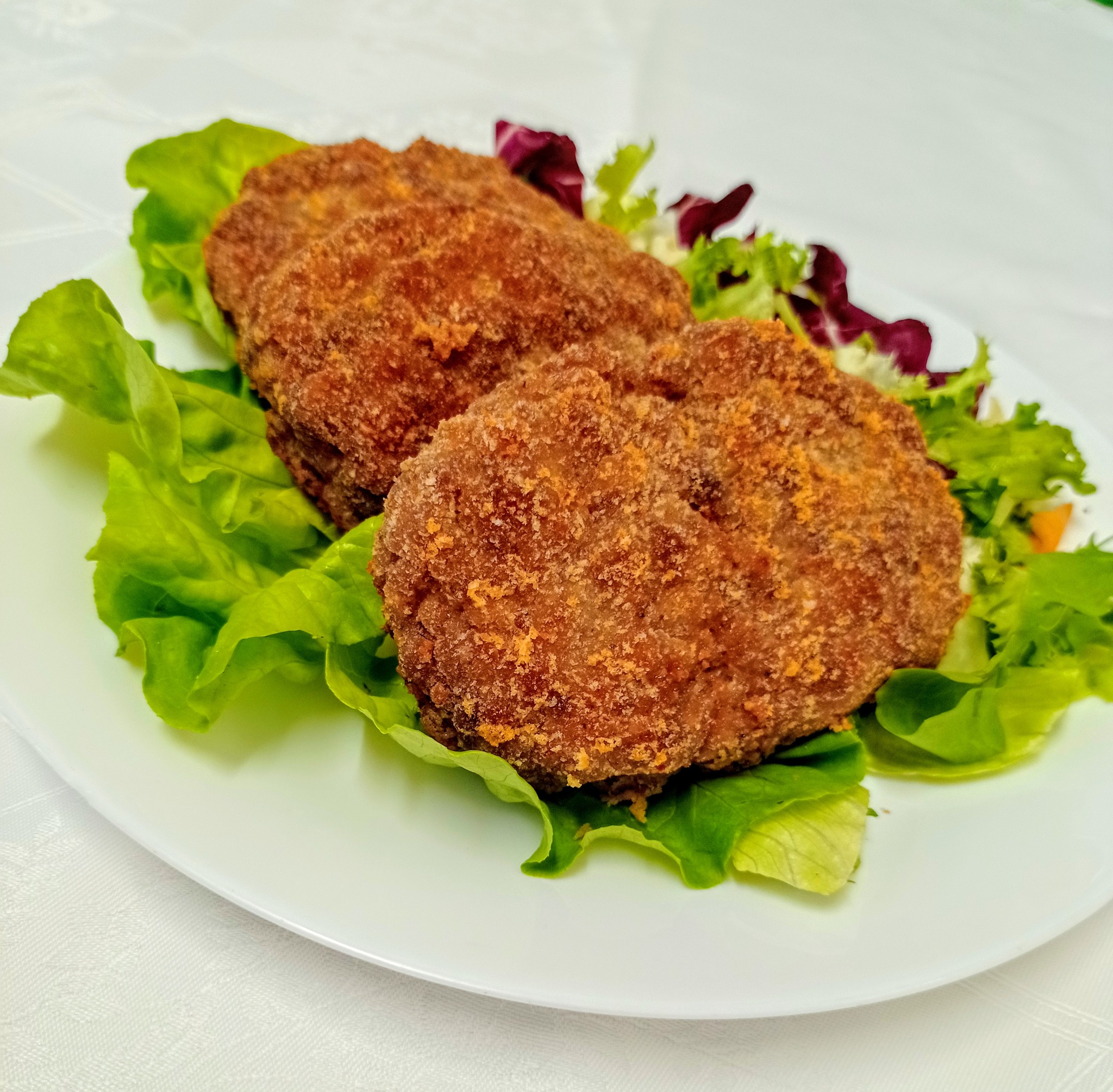 Trzy kotlety panierowane, ułożone na białym talerzu z zieloną i czerwoną sałatą, na tle białego obrusu.