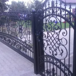 Czarna, metalowa brama wjazdowa z kutymi zdobieniami, widoczna z bliska na tle brukowanej ścieżki i domu z czerwonym dachem.