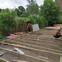 Dw&oacute;ch mężczyzn bez koszulek buduje drewniany taras na gruncie, widoczne elementy konstrukcyjne i narzędzia, w tle zieleń i dom.