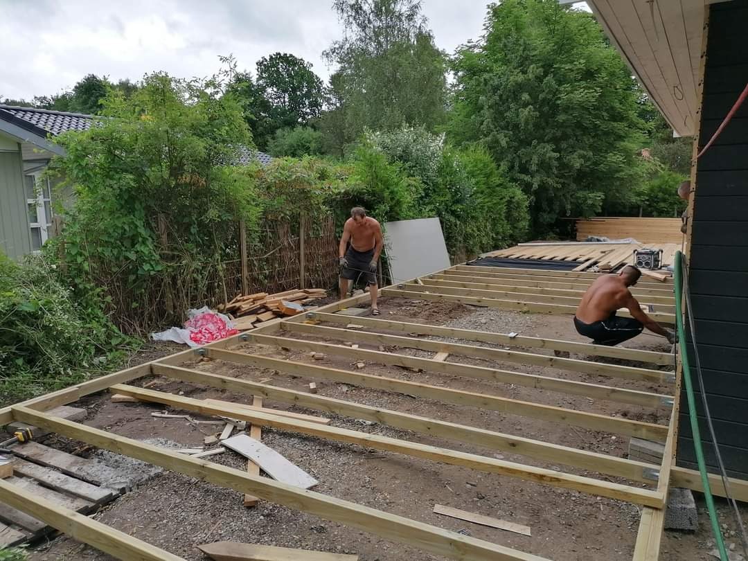 Dwóch mężczyzn bez koszulek buduje drewniany taras na gruncie, widoczne elementy konstrukcyjne i narzędzia, w tle zieleń i dom.