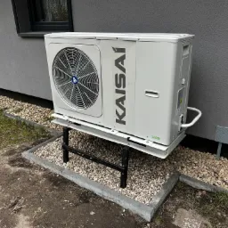 Zewnętrzna jednostka klimatyzacyjna marki Kaisai zamontowana na metalowym stelażu, umieszczona na kamiennym podłożu przy szarej ścianie budynku z widocznym oknem.