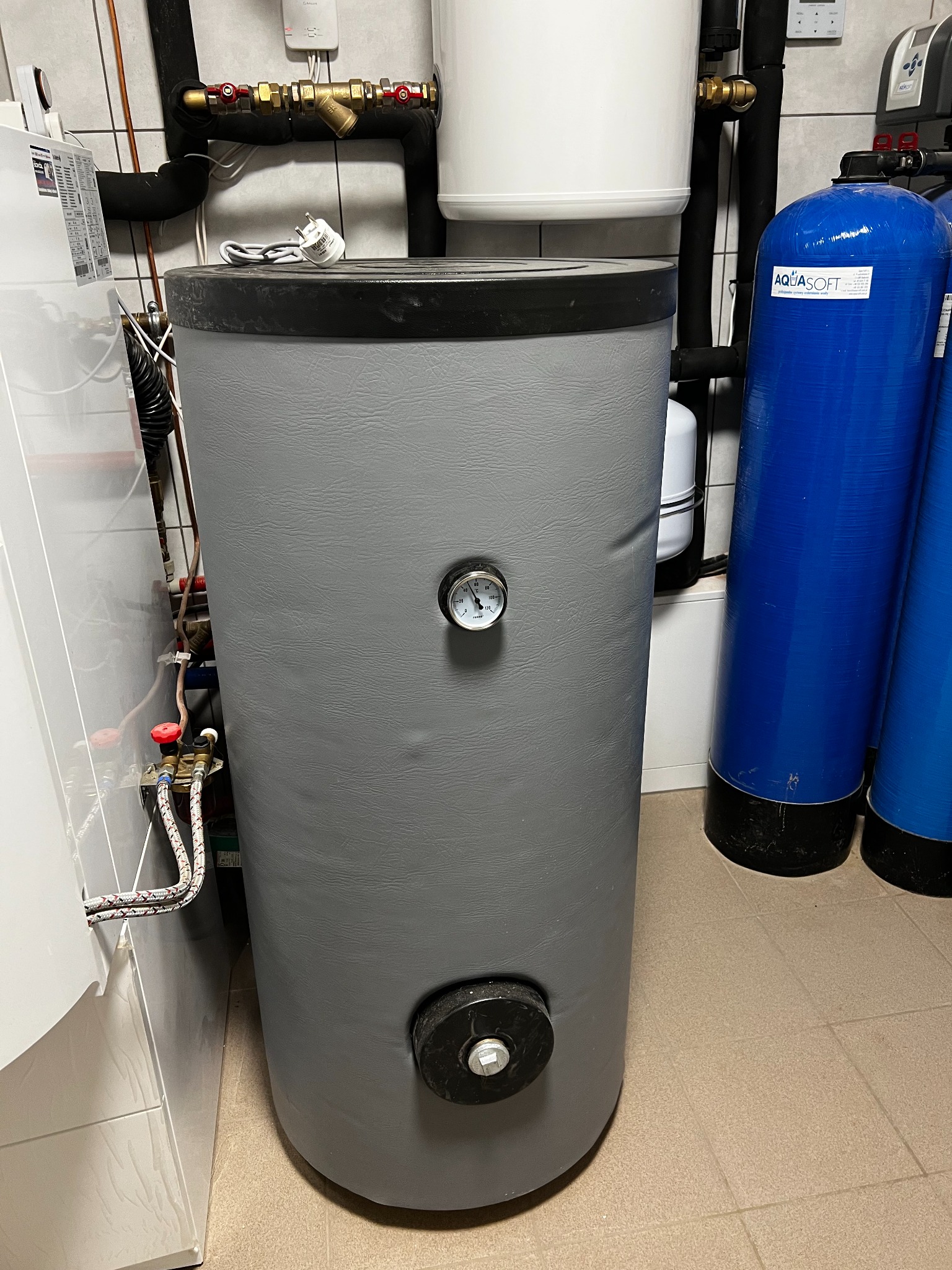 Szary, pionowy zbiornik buforowy z termometrem na środku, widoczne rury, zawory, biały bojler i niebieskie filtry wody w kotłowni wyłożonej kafelkami.