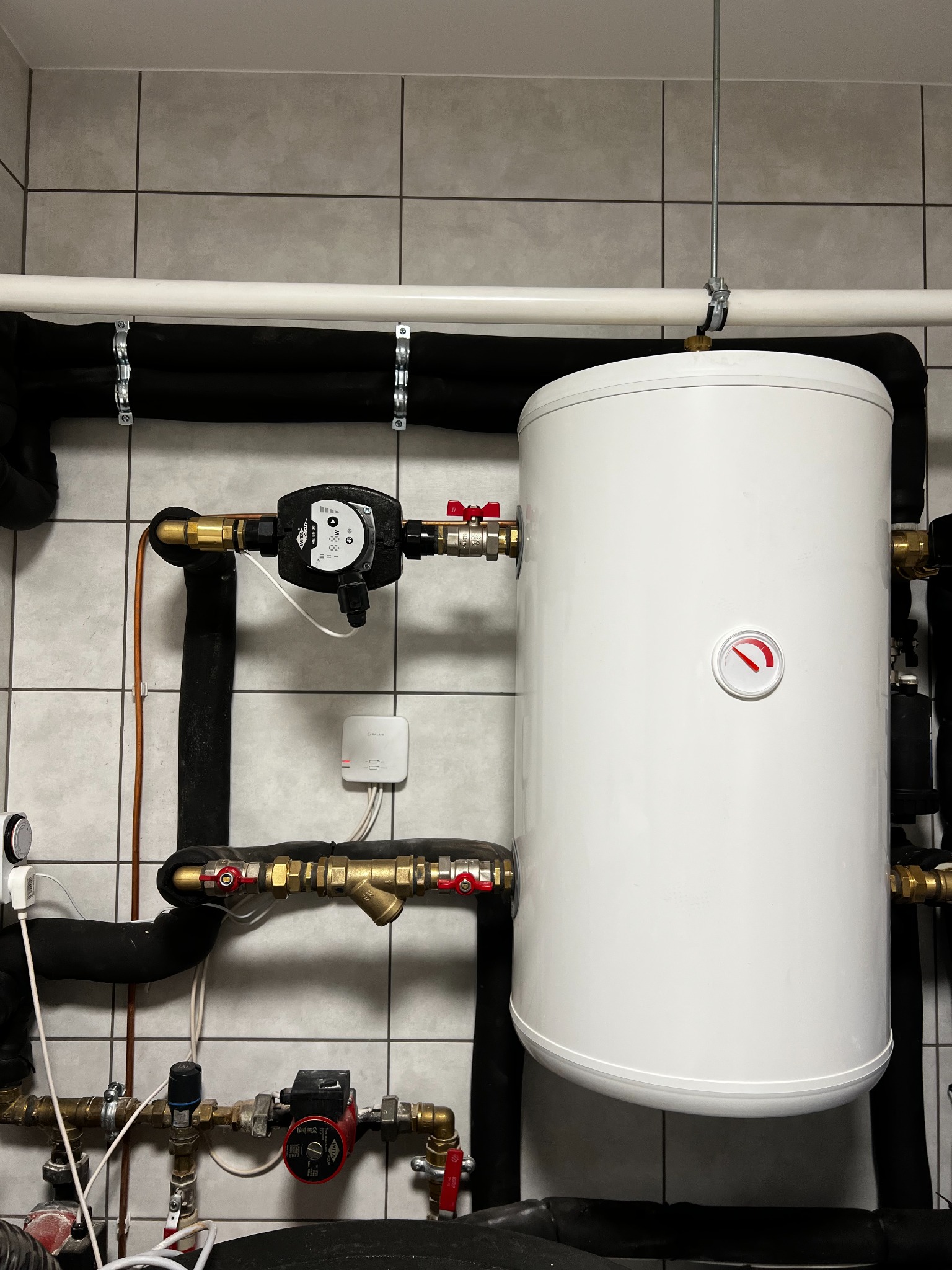 Biały zbiornik buforowy ciepła z widocznym termometrem, podłączony do instalacji z rur miedzianych i czarnych, izolowanych, na tle szarej, kwadratowej glazury.