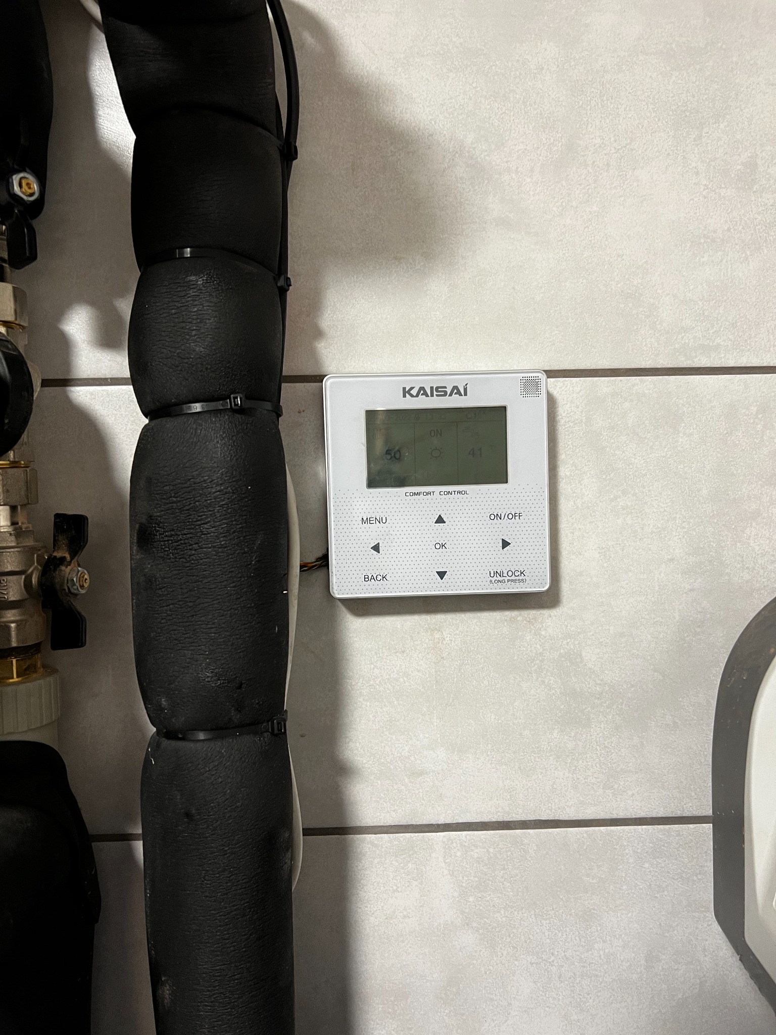 Sterownik pompy ciepła marki Kaisai zamontowany na kafelkowanej ścianie, obok zaizolowane rury instalacji grzewczej.