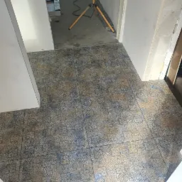 Ułożona podłoga z płytek w odcieniach niebieskiego i beżu z dekoracyjnym wzorem, widoczne fragmenty ścian i lampa budowlana w tle.