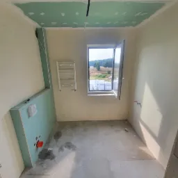 Łazienka w trakcie remontu: widoczne płyty gipsowo-kartonowe na suficie i ścianie, grzejnik drabinkowy, okno z widokiem na krajobraz, surowa betonowa posadzka, instalacja wodna.