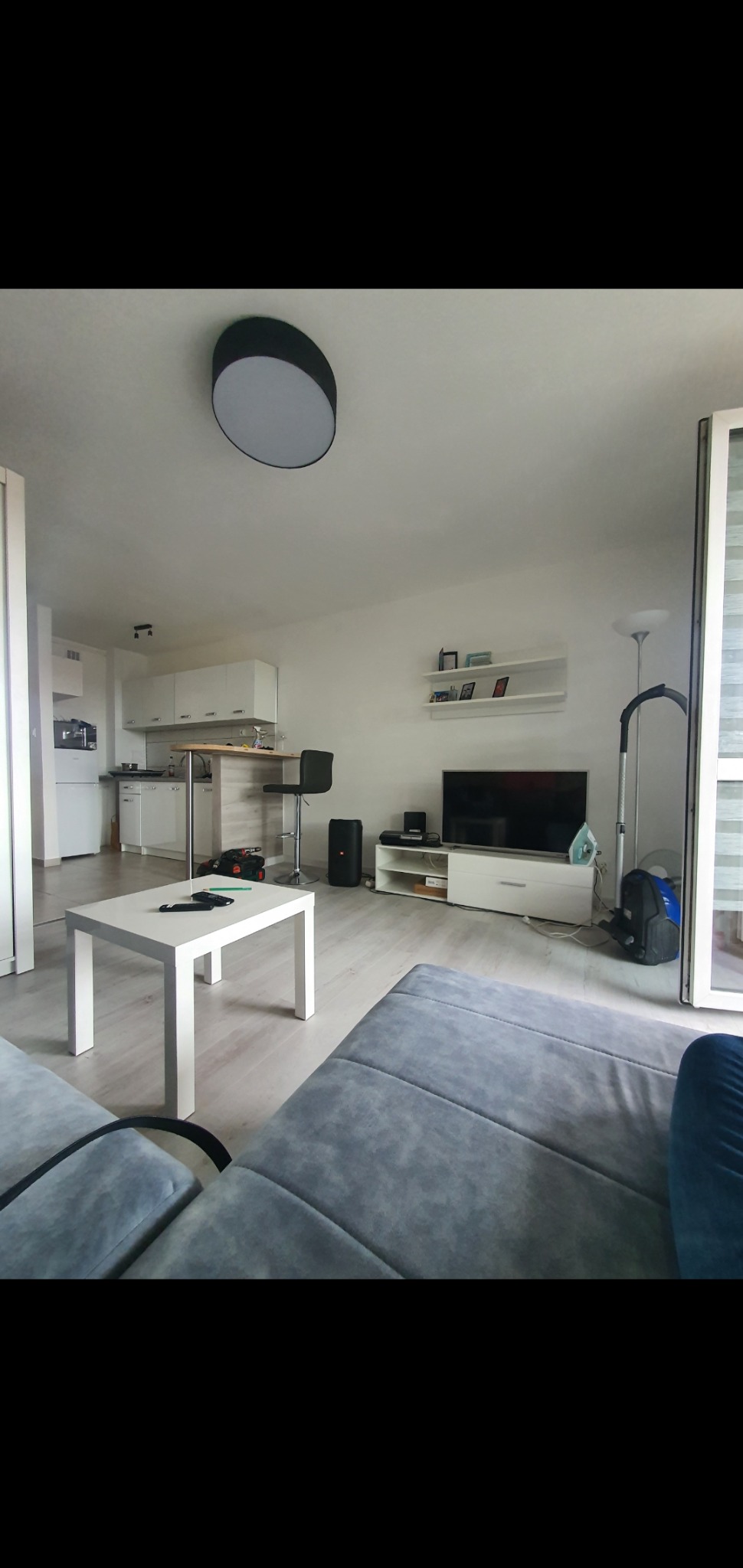 Jasne wnętrze mieszkania po remoncie: widok na salon z szarą sofą, biały stolik kawowy, aneks kuchenny z barkiem i krzesłem barowym, telewizor na białej szafce RTV, białe ściany i sufit z okrągłą...