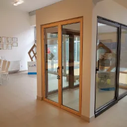 Ekspozycja w salonie: przesuwne drzwi szklane z drewnianą ramą oraz drzwi szklane z ciemną ramą, widoczne certyfikaty na ścianie w tle.