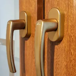 Dwa złote uchwyty okienne marki Roto zamontowane na drewnopodobnej ramie okiennej, widoczne delikatne odblaski na powierzchni uchwytów.