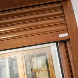 Drewnopodobna roleta zewnętrzna z widocznym oknem w tle, zamknięta, z logo producenta w prawym górnym rogu.