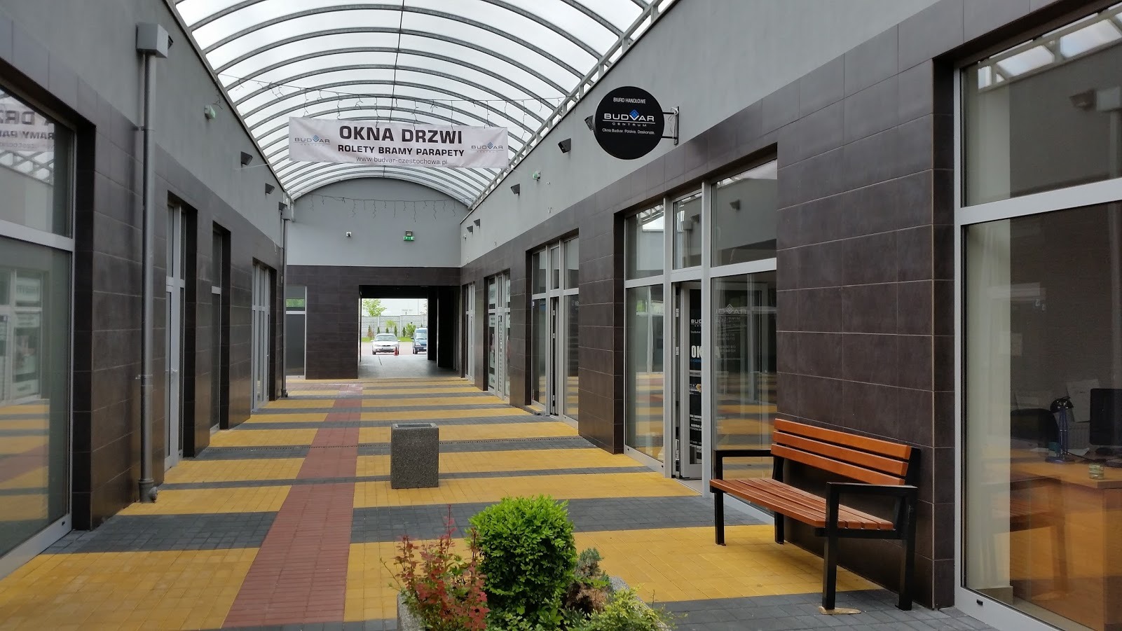 Widok na pasaż handlowy we Wrocławiu z witrynami salonu oferującego okna, drzwi, rolety i bramy, oświetlony naturalnym światłem przez przeszklony dach.