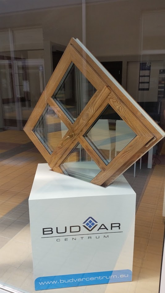 Przekrój okna z drewnopodobną ramą na białym postumencie z logo Budvar Centrum, prezentowany wewnątrz budynku z widocznym wnętrzem biura.