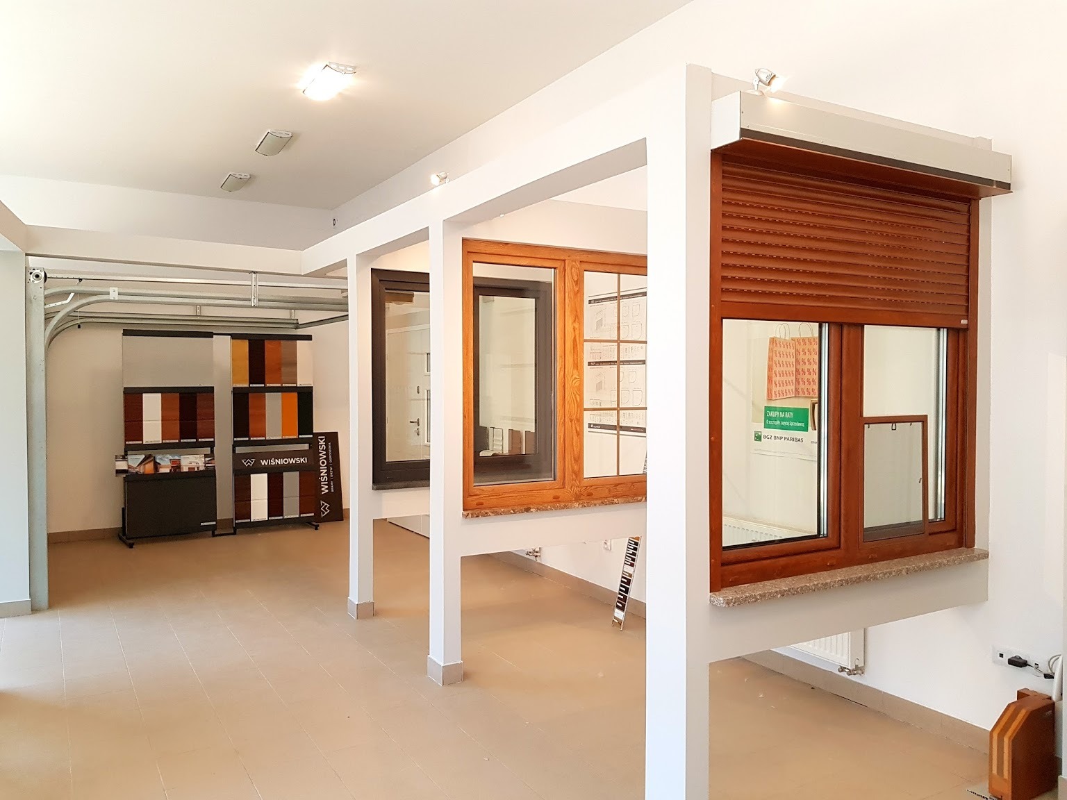 Ekspozycja w salonie: okno z brązową roletą zewnętrzną, okno drewniane i okno w ciemnej ramie, obok wzorniki kolorów.