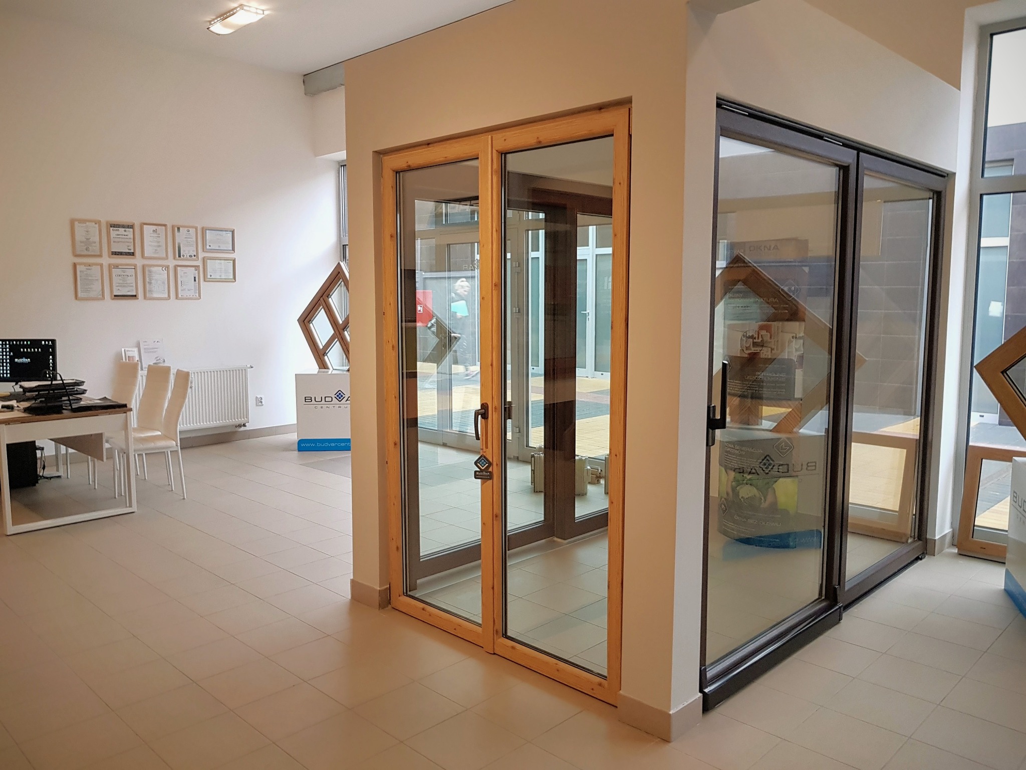 Ekspozycja w salonie: przesuwne drzwi szklane z drewnianą ramą oraz drzwi szklane z ciemną ramą, widoczne certyfikaty na ścianie w tle.