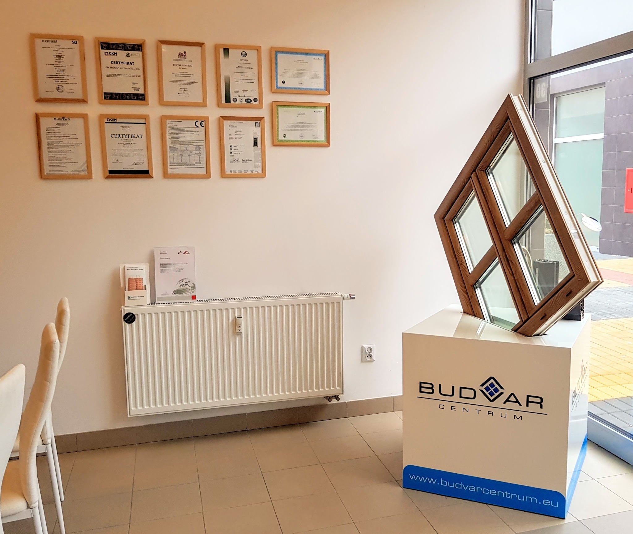 Prezentacja okna o nietypowym kształcie z brązową ramą na białym postumencie z logo Budvar Centrum, na tle ściany z certyfikatami i grzejnikiem.