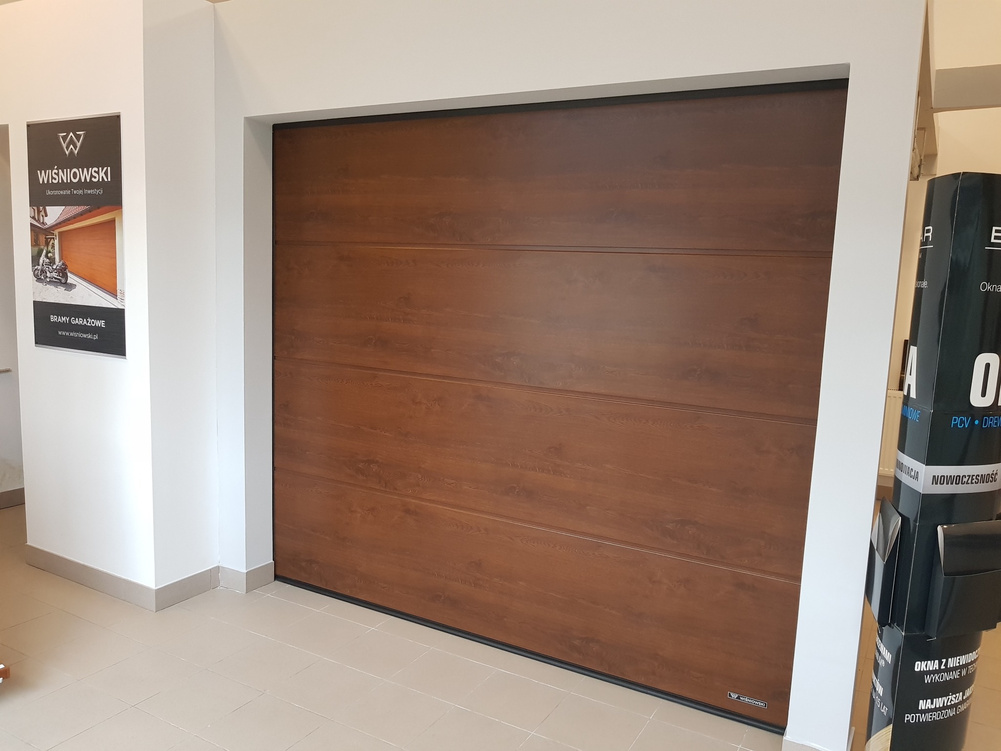Segmentowa brama garażowa Wiśniowski w kolorze drewna, prezentowana w salonie, z widocznym logo producenta na ścianie i materiałami reklamowymi obok.
