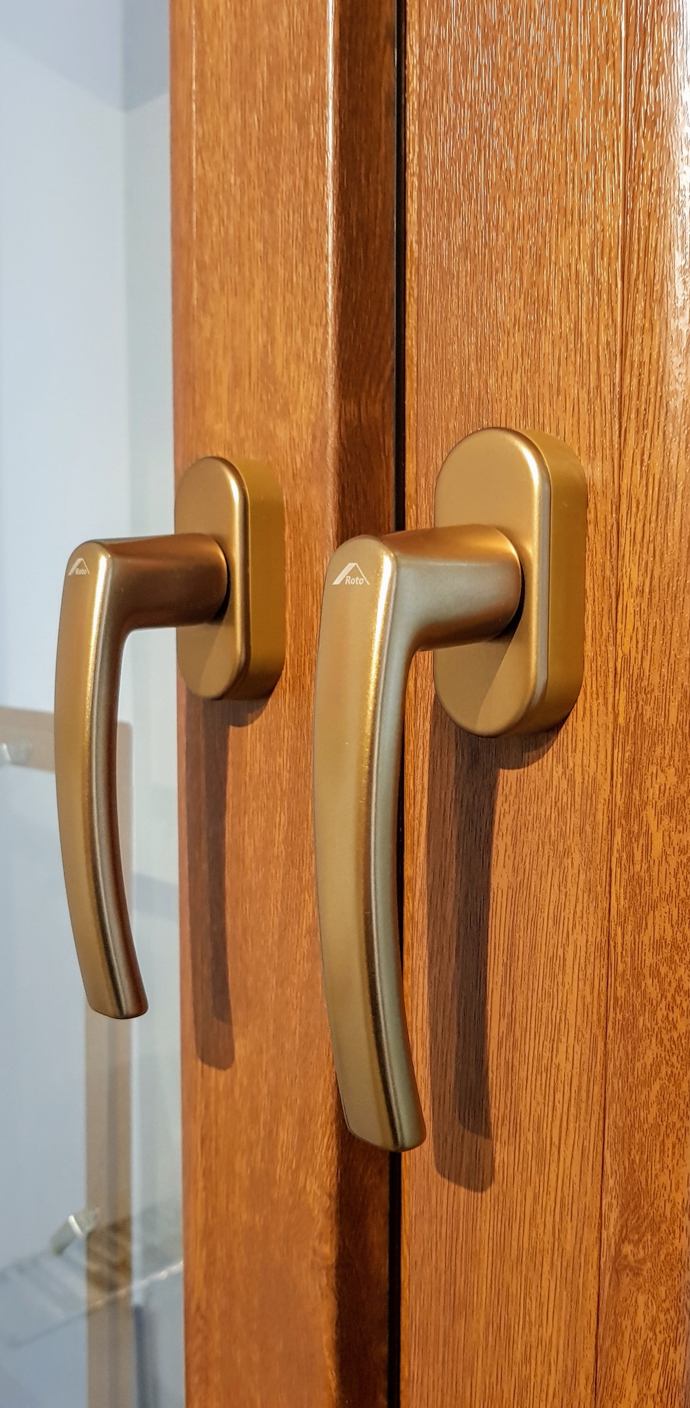 Dwa złote uchwyty okienne marki Roto zamontowane na drewnopodobnej ramie okiennej, widoczne delikatne odblaski na powierzchni uchwytów.