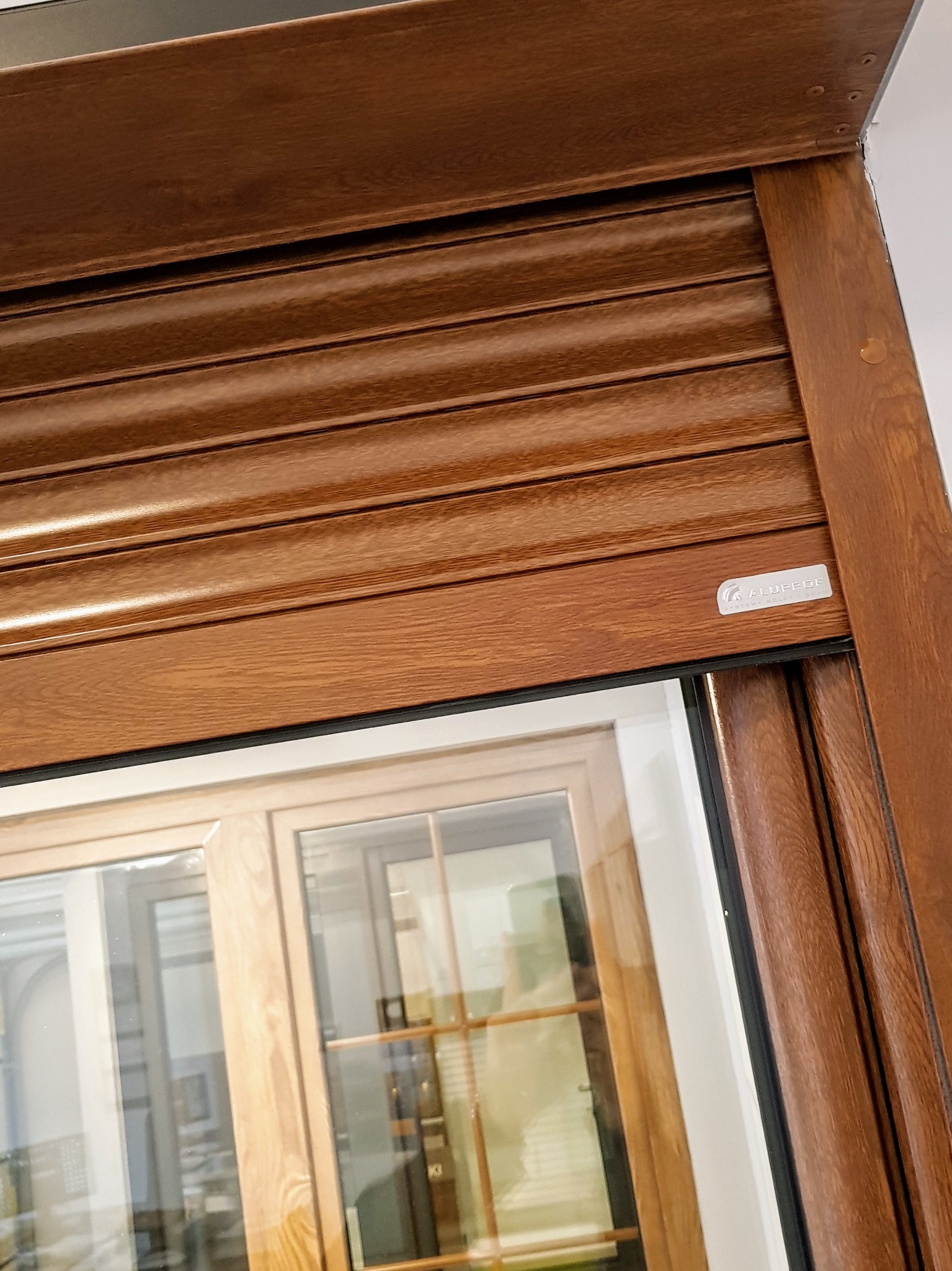 Drewnopodobna roleta zewnętrzna z widocznym oknem w tle, zamknięta, z logo producenta w prawym górnym rogu.