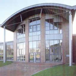 Budynek z cegły z dużymi, szarymi oknami PCV i zaokrąglonym dachem, widoczne kolumny wspierające dach.