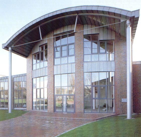 Budynek z cegły z dużymi, szarymi oknami PCV i zaokrąglonym dachem, widoczne kolumny wspierające dach.