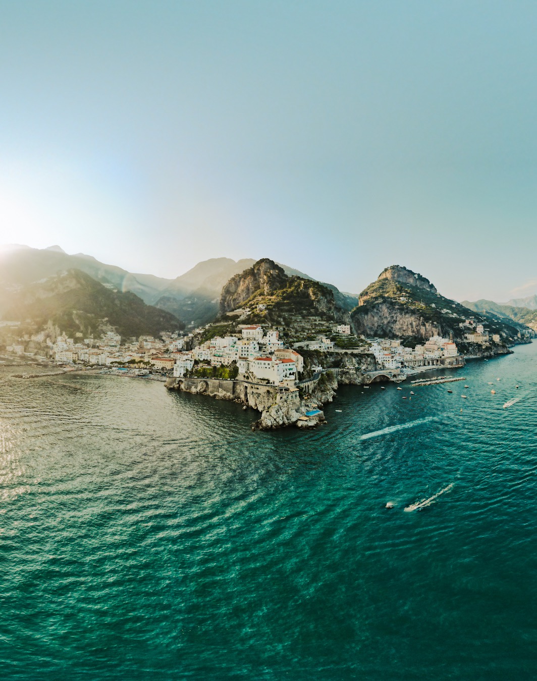 Panoramiczny widok z lotu ptaka na malownicze miasteczko położone na skalistym wybrzeżu, otoczone turkusowym morzem i górami w tle, z promieniami słońca przebijającymi się przez chmury.