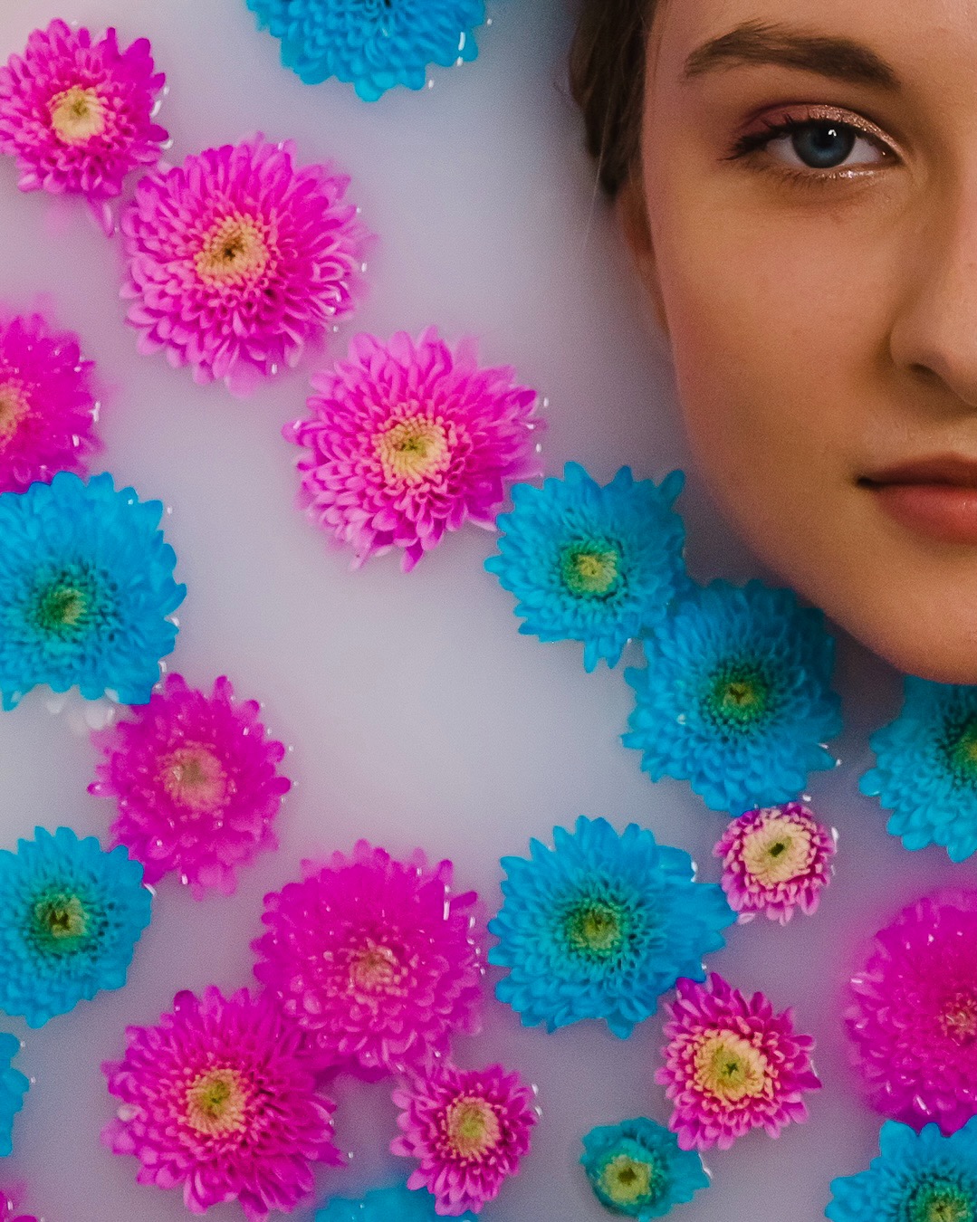 Portret kobiety zanurzonej w mlecznej kąpieli z pływającymi różowymi i turkusowymi chryzantemami, z widocznym makijażem oczu i delikatnymi ustami.