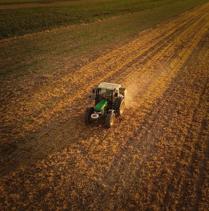 Zielony traktor John Deere na polu z widocznymi śladami opon, sfotografowany z góry w ciepłym świetle zachodzącego słońca.