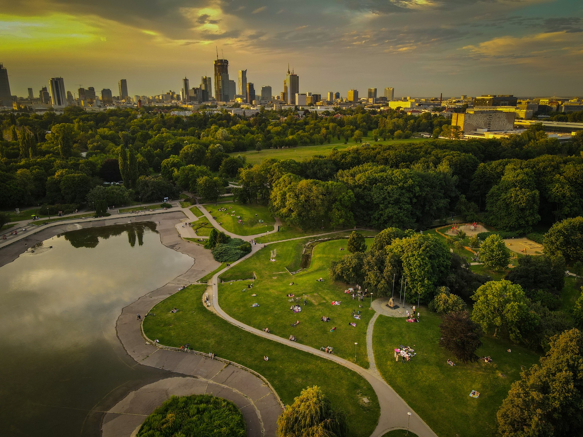 Panorama Warszawy z lotu ptaka, park z jeziorem i wypoczywającymi ludźmi w promieniach zachodzącego słońca.